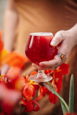 Hand with silver rings holds a stemmed glass of deep red cocktail beside vibrant orange and red flowers.