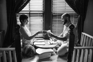 Black and white photo of a couple sitting on a wooden floor by a window, clinking mugs over a small table with papers and books.