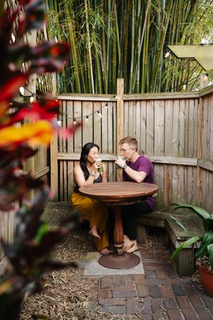 Two people sit at a round wooden table in a cozy backyard patio with bamboo, string lights and a wooden fence, sharing drinks and smiling.