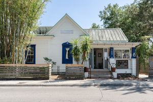 White wooden cottage with blue shutters, a metal roof, front porch, and wooden fence, shaded by tall bamboo and trees on a sunny street.