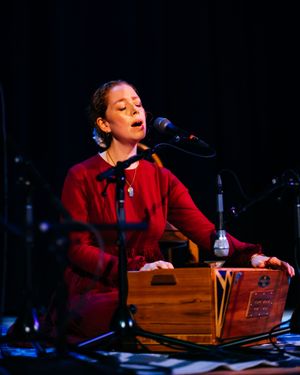 Woman in a red dress sings into a microphone while playing a wooden harmonium on a dimly lit stage during a live concert performance.
