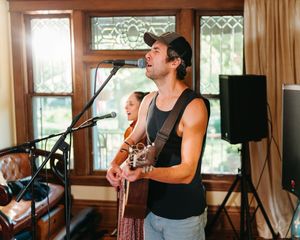 Man in a backwards cap and black tank top plays acoustic guitar and sings into a microphone during an intimate indoor live music session.