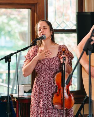 A woman in a floral dress sings into a microphone with her hand on her chest, holding a violin during a live indoor performance.