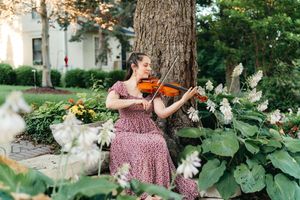 Woman in a floral dress playing a violin outdoors beside a large tree, surrounded by white hydrangeas and green hostas in a garden.