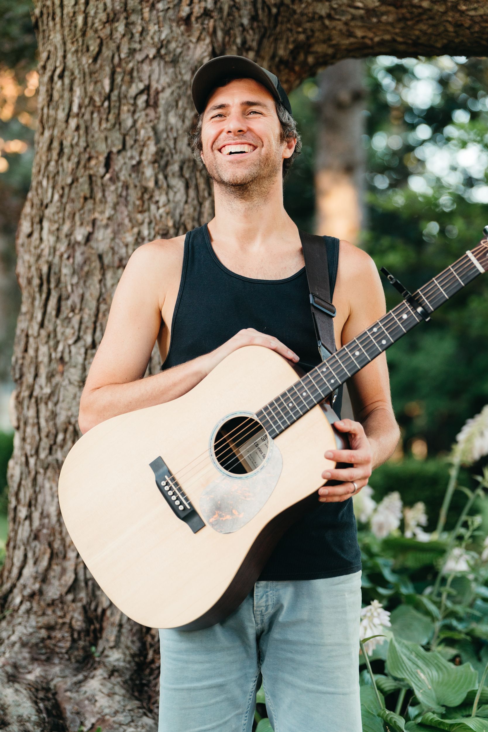 Smiling man in a black tank top and cap holds an acoustic guitar while leaning against a large tree beside blooming garden flowers.