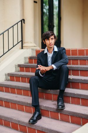 Young man in a dark suit and black dress shoes sits on red brick steps outside a columned building, hands clasped, looking at the camera.