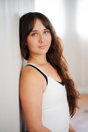 Young woman with long wavy brown hair and light eyes leans against a white wall in a white tank top with black straps.