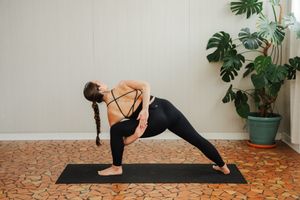 Woman with a long braid practices a revolved side angle yoga pose on a black mat beside a potted monstera plant in a bright studio.