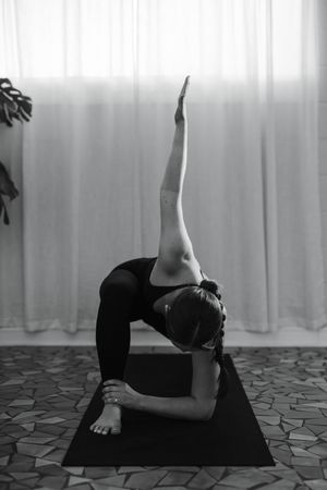 Black and white photo of a woman in a yoga twist pose on a mat, one arm reaching upward, with sheer curtains and a mosaic floor behind her.