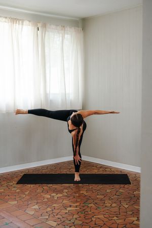 Woman in black activewear performs a standing split yoga pose on a black mat beside sheer curtains in a bright studio with a tiled floor.
