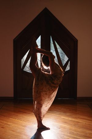 Silhouetted woman in a flowing dress performs a standing bow yoga pose on a polished wood floor before a pentagon-shaped glass door.