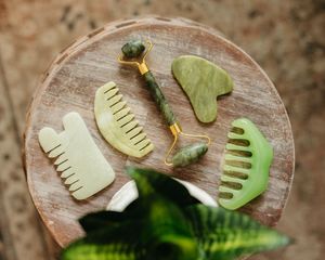 Set of green jade gua sha tools and a jade facial roller with gold handle arranged on a round wooden table beside a leafy plant.