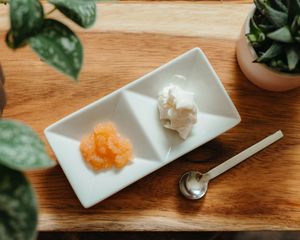 Divided white ceramic dish holding orange fruit puree and whipped cream beside a small spoon on a wooden board with green plants.