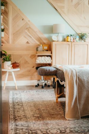 Cozy spa treatment room with a massage table, herringbone wood accent wall, vintage rug, towel shelf, and potted plants.