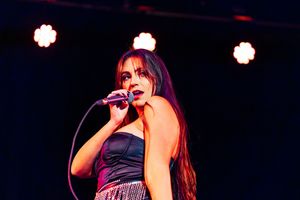 Woman with long dark hair sings into a microphone on stage in a dark corset top and sparkly skirt under glowing round stage lights.