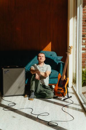 Man sits on the floor in sunlight with closed eyes, leaning against a teal sofa beside an orange bass guitar and a Fender amplifier.