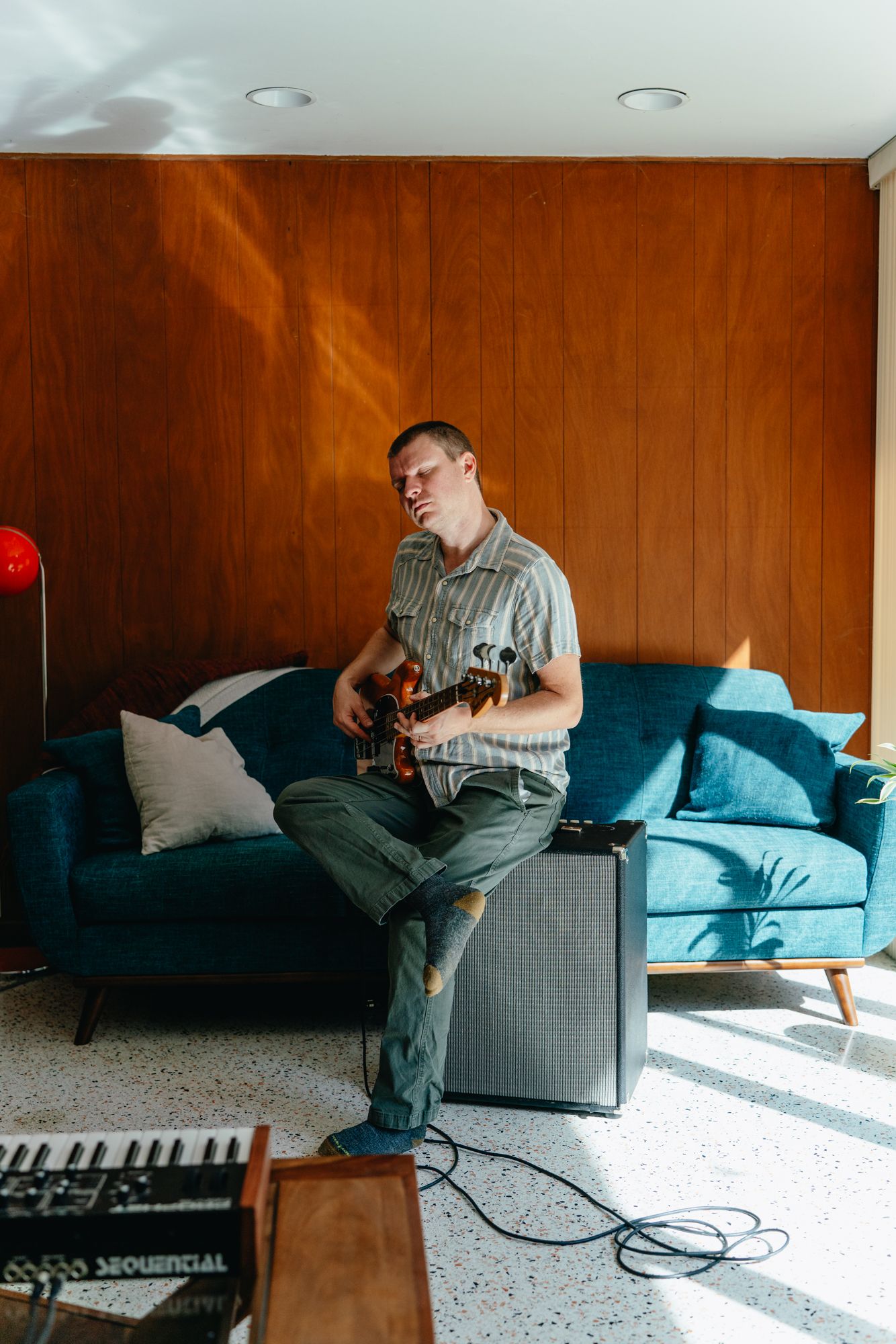 Man in a striped shirt sits on an amplifier playing electric guitar in a mid-century living room with wood paneling and a teal sofa.