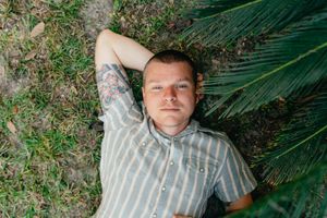 Man with a tattooed arm in a striped short-sleeve shirt lies on green grass beside palm fronds, resting his head on one hand.