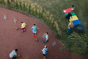 Aerial view of children and adults playing on a red rubber playground beside a colorful hopscotch mat, trampoline and tall grass.
