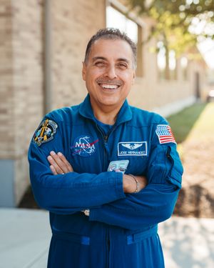 Smiling man with arms crossed wears a blue NASA flight suit with name tag and American flag patch, standing outside a brick building.
