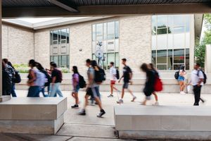 Blurred students with backpacks walk briskly along a sunlit brick campus building with large windows and concrete benches.