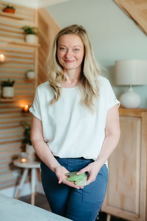 Smiling woman with long blonde hair in a white blouse and blue pants holds green gua sha stones in a cozy spa room with candles and plants.