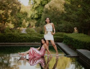 Two women in elegant dresses pose by a reflective garden pool, one standing in white and one seated in pink, surrounded by lush green hedges