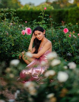 Woman with long dark hair in a shimmering pink dress sits among blooming pink and white roses in a lush garden.