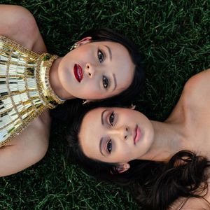Two women with dark hair lie on green grass in an overhead shot, one wearing a gold embellished top and bold red lipstick.