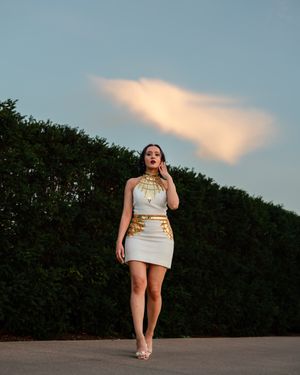 Woman in a white halter mini dress with gold embellishments poses on pavement before a tall green hedge and a glowing pink cloud.
