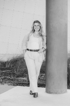 Smiling woman in glasses, striped open shirt, and wide-leg pants leans against a concrete column in a black and white outdoor portrait.