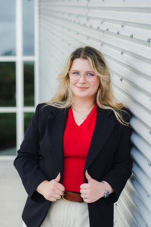 Smiling woman with long blonde hair and glasses wears a black blazer and red top while leaning against a corrugated metal wall.