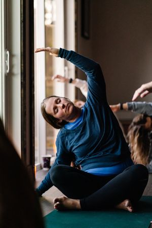 Woman sitting cross-legged on a green yoga mat performs a side stretch with one arm raised during a group yoga class by a sunlit window.