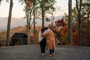 Couple embracing on a paved overlook surrounded by vibrant autumn foliage, with a cabin and misty mountains in the background.