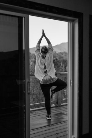 Woman practicing tree pose yoga on a wooden deck with hands in prayer position, overlooking forested mountains in black and white.