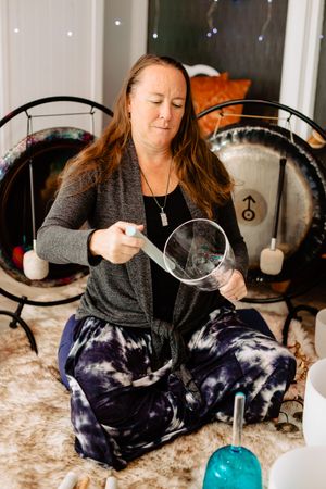 Woman sitting cross-legged on a fur rug plays a crystal singing bowl with a mallet during a sound healing session, surrounded by gongs.