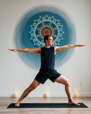 Man in black athletic wear practices Warrior II yoga pose on a black mat in front of a blue mandala wall mural, with candles nearby.