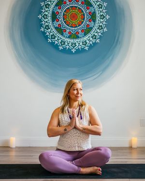 Woman with blonde hair sits cross-legged in prayer pose on a black yoga mat in front of a blue mandala mural, with candles beside her.