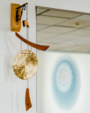 Hanging brass gong with curved wooden striker and mallet on a wall bracket beside a mirror reflecting a soft blue mandala in a calm studio.