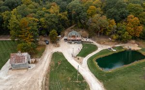 Aerial view of a rustic cabin and barn on a wooded property with a winding gravel driveway, green lawn, and a small pond in autumn.