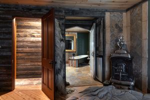 Rustic log cabin bathroom with a freestanding soaking tub, stone walls, reclaimed wood paneling, and a vintage cast iron wood stove.
