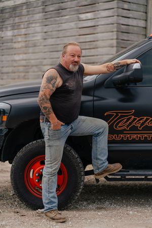 Man with tattooed arms and a gray beard stands with one boot on the step of a black pickup truck with red wheels beside a weathered wall.