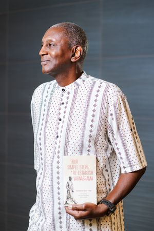 Smiling man in a patterned short-sleeved shirt looks to the side while holding a book titled Four Simple Steps to Re-establish Varnashrama.