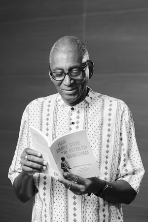 Smiling man in glasses and a patterned short-sleeve shirt reads a booklet titled Four Simple Steps to Re-establish Varnashrama.