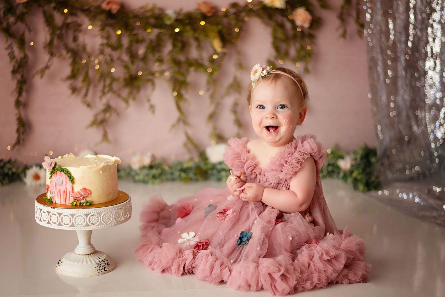 Smiling baby girl in a pink tulle ruffle dress and flower headband sits beside a pink floral smash cake at a first birthday photoshoot.