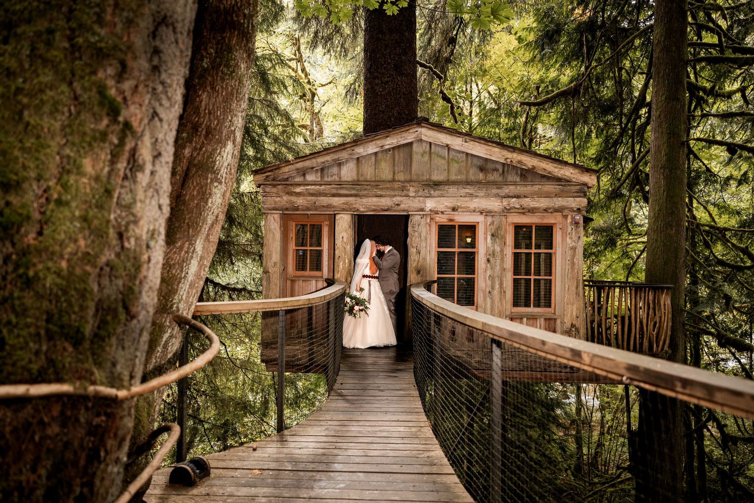 Bride and groom embrace in the doorway of a rustic treehouse cabin at the end of a wooden suspension bridge in a lush forest.