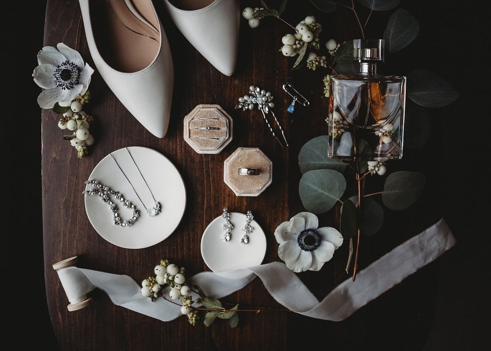 Flat lay of bridal accessories on a dark wooden table with ivory heels, pearl jewelry, two rings, perfume, white flowers and a satin ribbon.