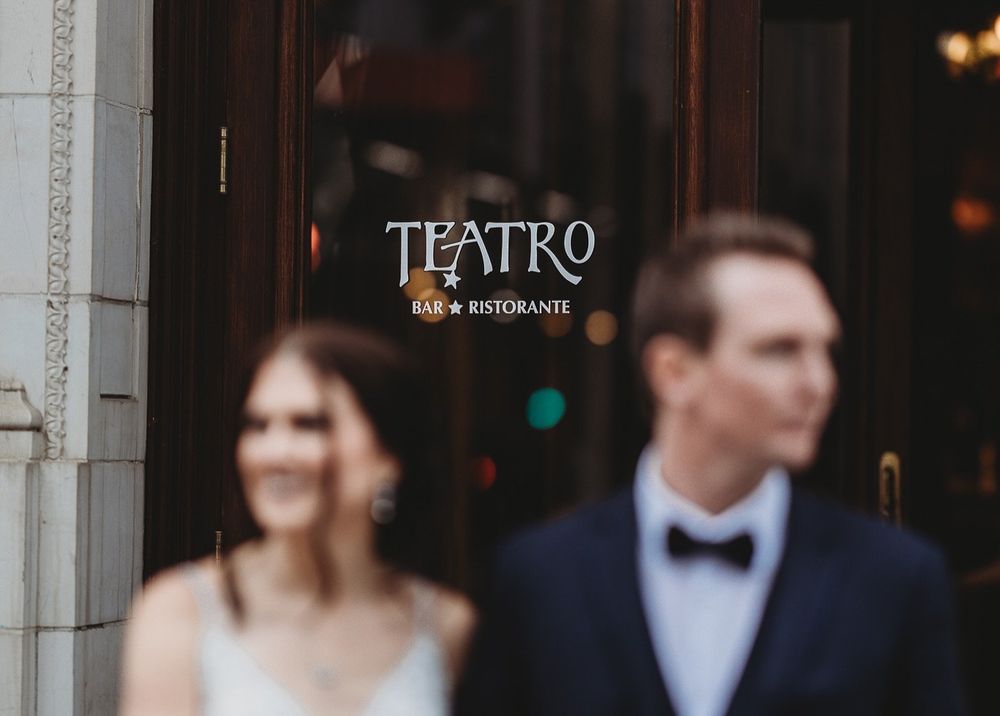 Blurred bride and groom in formal attire stand outside Teatro Bar Ristorante, whose elegant sign glows on the dark glass door behind them.