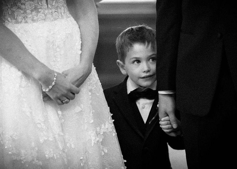 Young boy in a black tuxedo and bow tie peeks out between a bride in a lace wedding gown and a groom during a wedding ceremony.