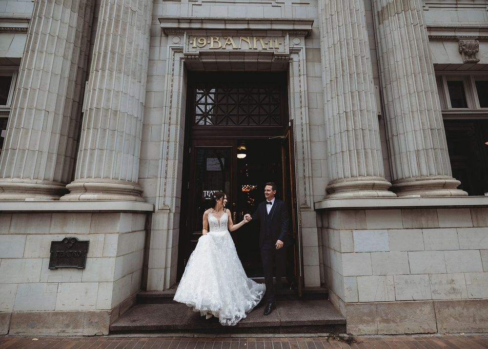 Bride in a white ball gown and groom in a dark suit stand hand in hand in the doorway of a grand stone building with tall columns.